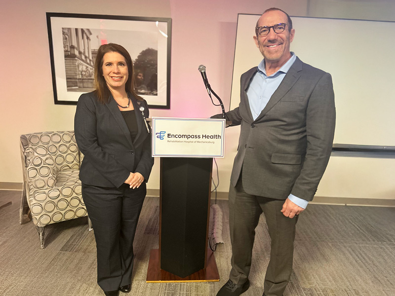 Two speakers stand at a podium in front of a large screen, presenting to an audience.