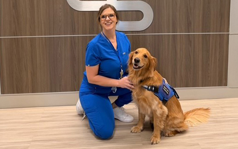 A woman in scrubs kneels beside a therapy dog, showing care and attention in a medical setting.