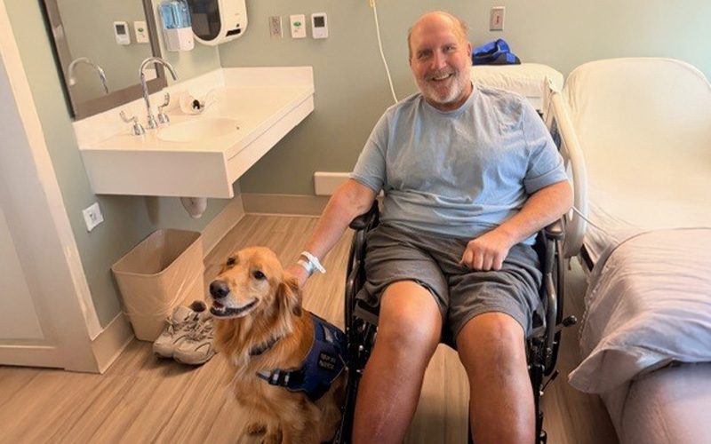A man in a wheelchair shares a hospital room with a therapy dog, symbolizing support and comfort in a medical setting.