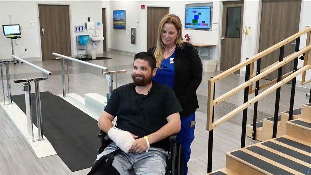 A man with a bandaged arm sits in a wheelchair, smiling, assisted by a nurse in blue scrubs in a rehabilitation facility with therapy equipment.