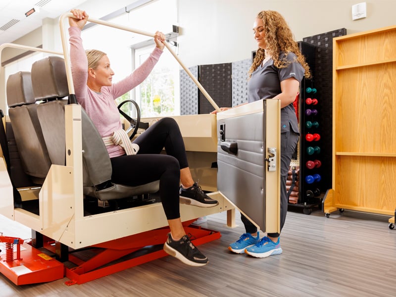 A woman practices transfer skills from a car simulator with assistance from a therapist in a rehabilitation center. The setting is supportive and encouraging.