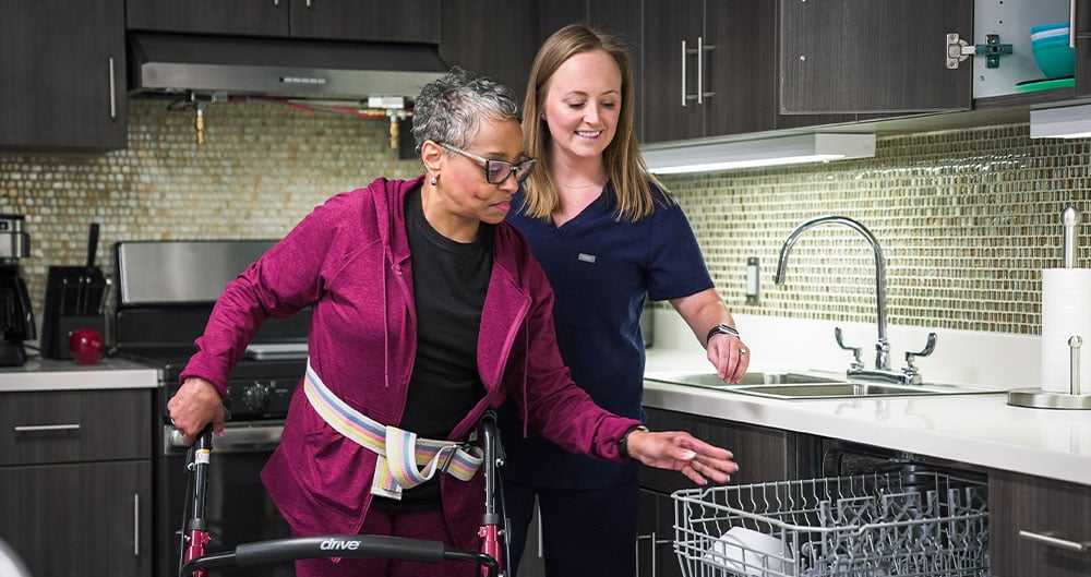 An occupational therapist helps an elderly woman using a walker load a dishwasher in a modern kitchen, conveying support, independence, and assistance.