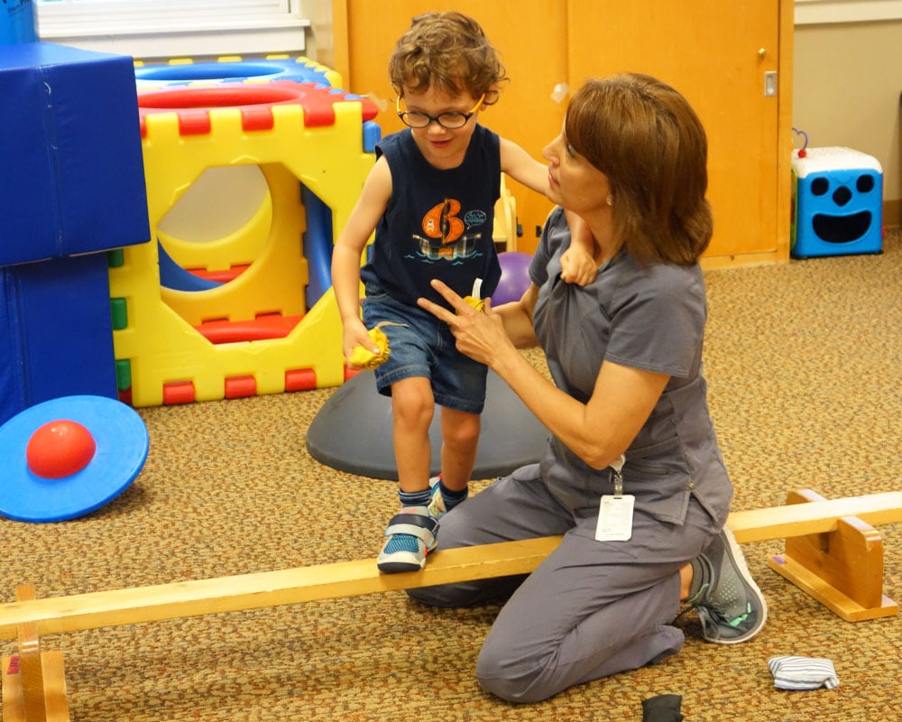 A child with glasses balances on a wooden beam, supported by a therapist kneeling beside him. The setting is colorful and playful, conveying encouragement.