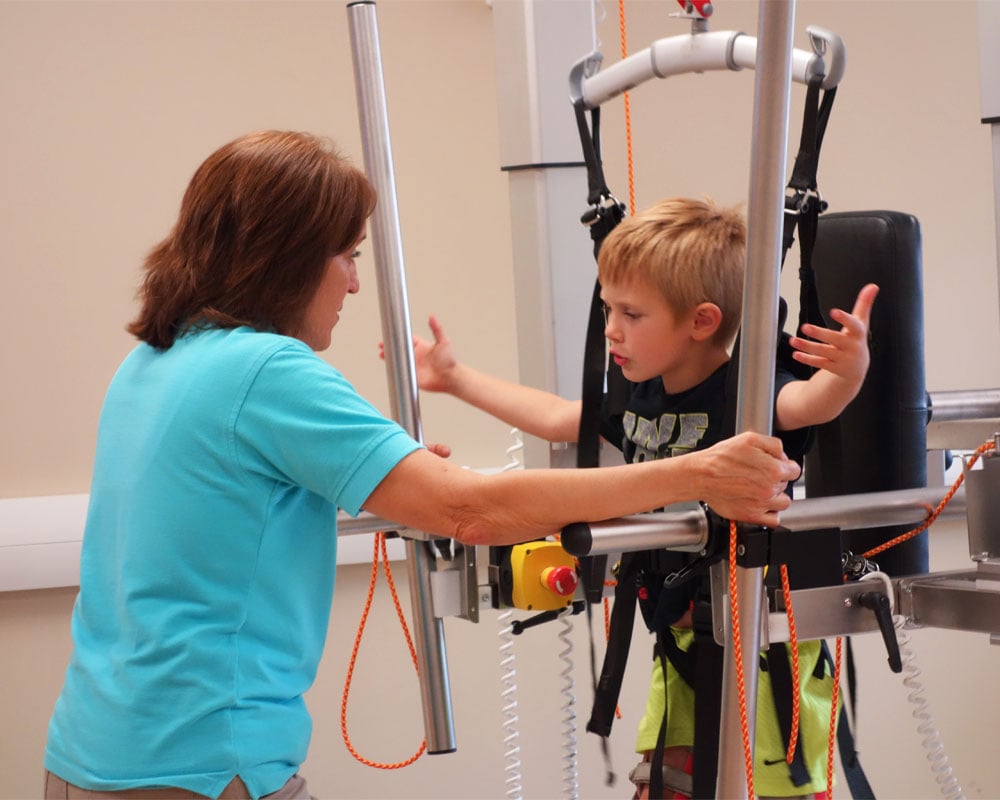 A therapist assists a young boy in a harness during a physical therapy session. The boy is focused and engaged, conveying determination and support.