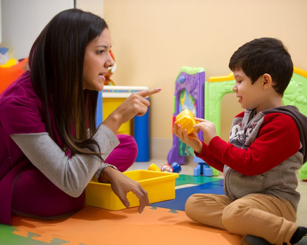 A woman and a young boy sit on a colorful play mat. The woman gestures as the boy holds a yellow toy, suggesting a learning moment in a playful setting.