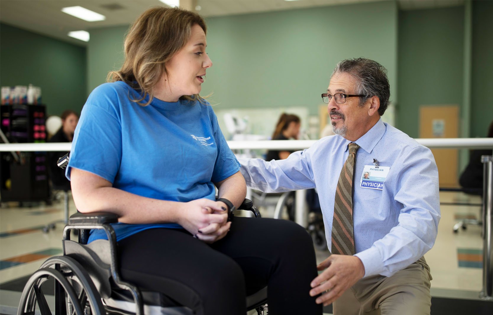 A physician speaks with a patient in a wheelchair during a rehabilitation session in a therapy gym.