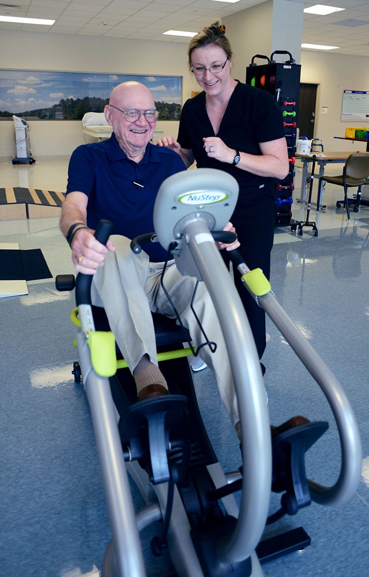 An elderly man smiles while using a NuStep exercise machine, assisted by a supportive woman in a bright, spacious rehab facility.