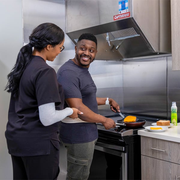 A man smiles while cooking at a stove, holding a pan with a cheese-topped dish. A woman in scrubs stands beside him, suggesting a friendly, casual atmosphere.