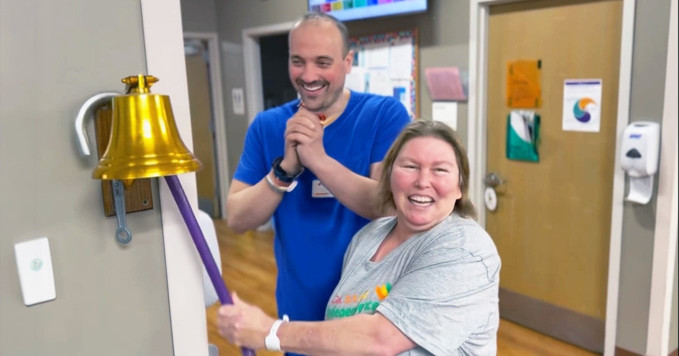 A joyful woman rings a brass bell mounted on a wall, celebrating a milestone. A smiling man in blue scrubs claps supportively behind her.