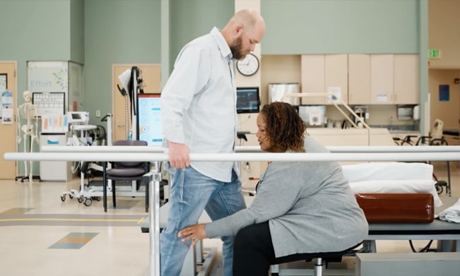 A man in a white shirt and jeans practices standing with assistance from a therapist in a gray sweater in a rehabilitation center equipped with parallel bars.