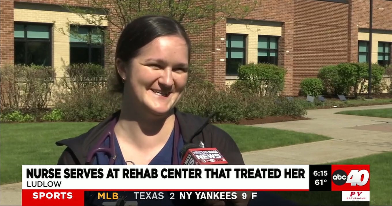A smiling nurse stands outside a brick rehab center with greenery. She's speaking to a microphone, conveying warmth and positivity. News banner text reads: Nurse serves at rehab center that treated her.