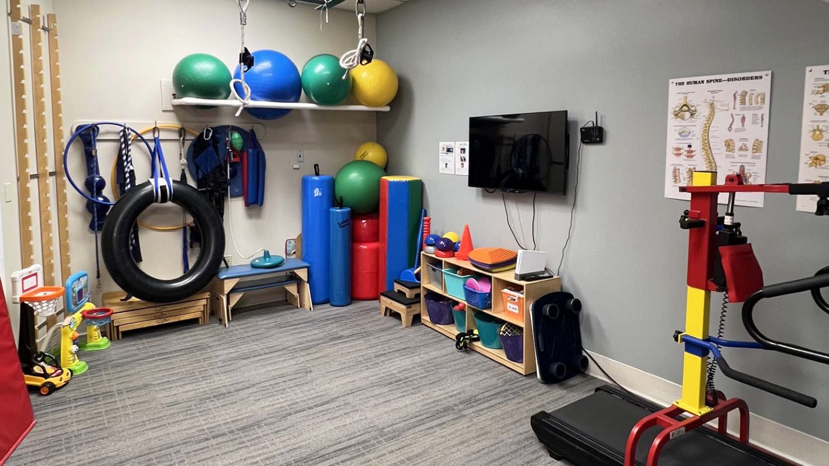 A therapy room with exercise equipment: colorful therapy balls, a tire swing, climbing wall elements, gymnastics rings, and a small trampoline. The space is organized and inviting.