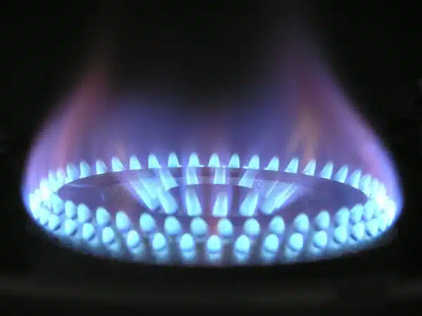 A close-up shot of a blue gas flame emanating from a circular burner with multiple small holes. The flame is bright blue at its base and transitions to a lighter blue at the tip. The background is dark and out of focus.