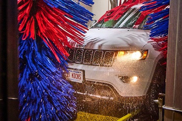 A white Jeep SUV is being washed with large red and blue brushes. The car's headlights are on, and water is spraying. A license plate reading 