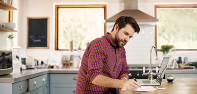 man writes on paper with laptop open in well furnished kitchen