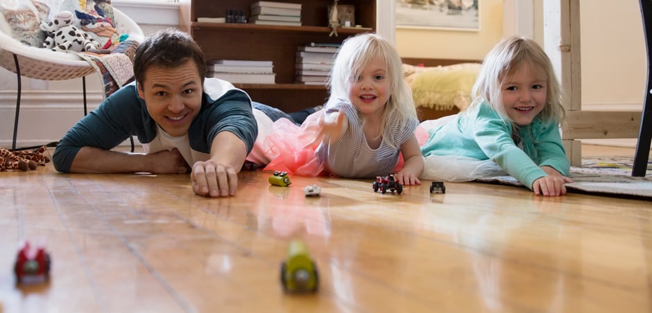 father and daughters play with toy cars on hardwood floor