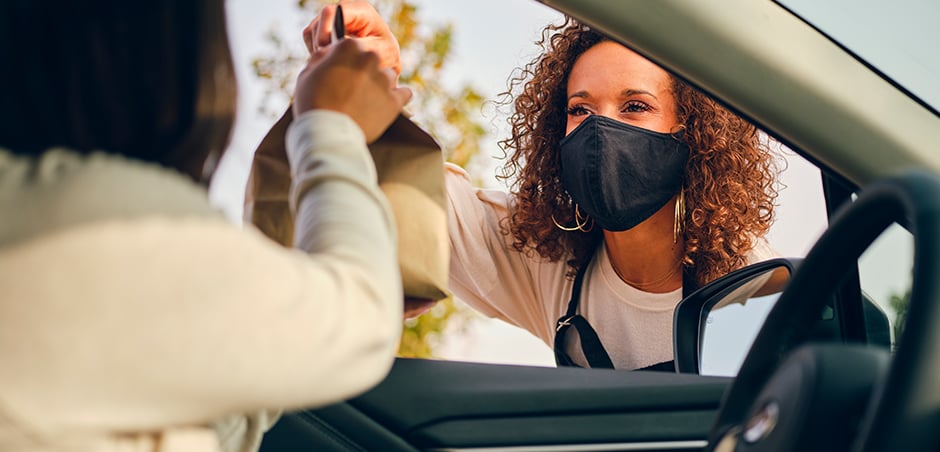 woman wearing mask hands brown paper bag to person in driver's seat of car