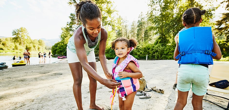 mother assists young daughter in putting on her life jacket, while older child looks ahead.