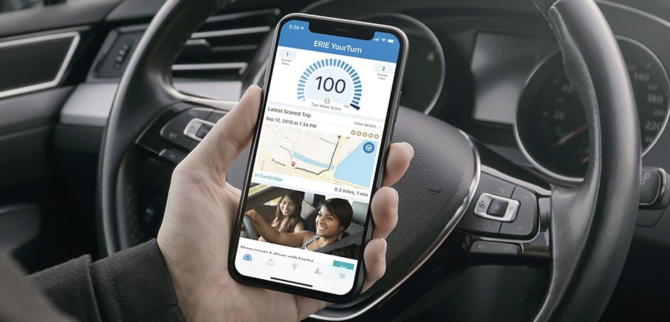 Hand holds a smartphone in front of a car steering wheel, with ERIE's YourTurn app showing on screen.