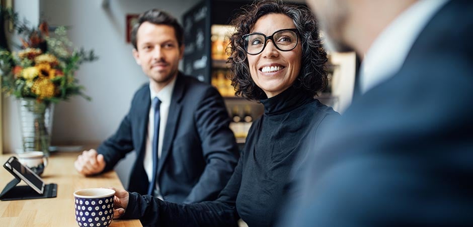 A man and woman sit at a coffee shop bar in business formal attire. A third man in a business suit is at front of frame, slightly out of focus.