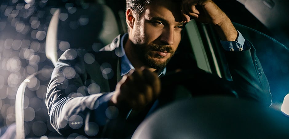 Professionally dressed man looks sleepy and rests his head on his hand while behind the wheel of a vehicle.