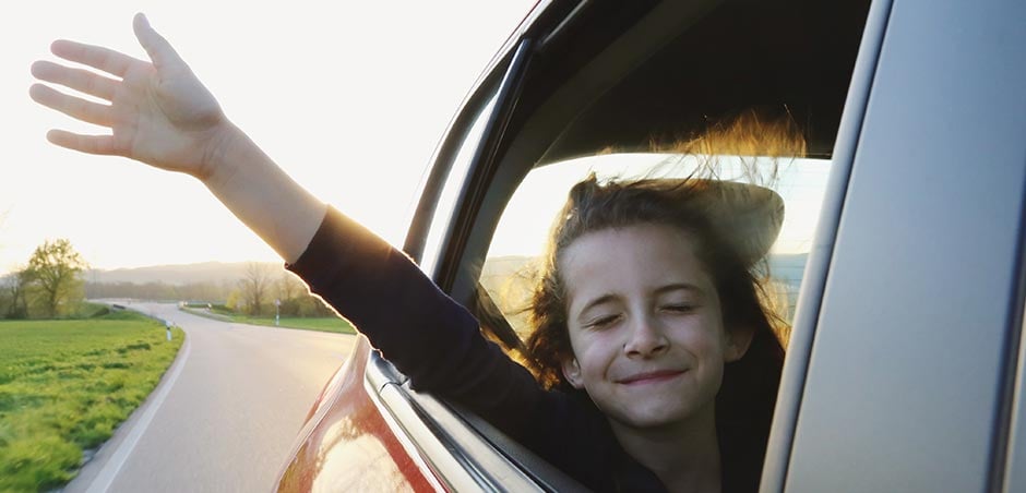 Child holds her hand out of the rear window of a moving car on a scenic country road.