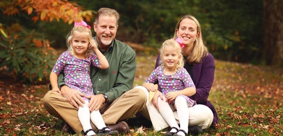 Portrait of the Baker family, with four people seated on the grass. The two young daughters sit on their mother and father's laps.