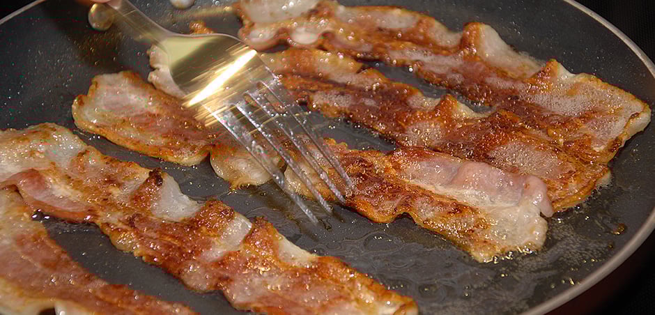 a fork poking bacon strips in a frying pan