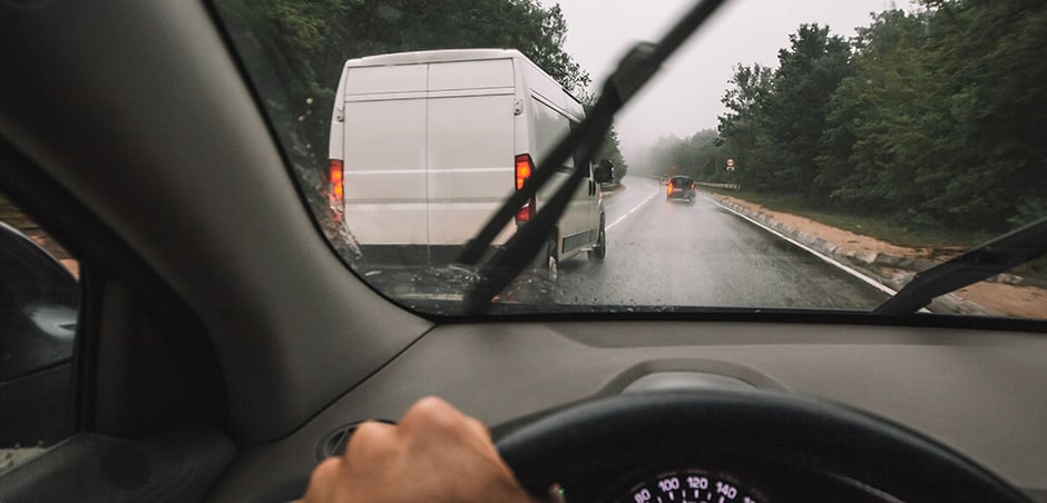 person with one hand on the steering wheel is driving down the highway with their windshield wipers on in the right lane. a white van is ahead in the left lane 