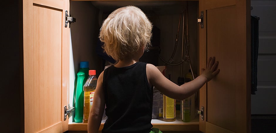 A young child stands in front of an open cabinet. Bottles of various cleaning products are visible inside the cabinet.