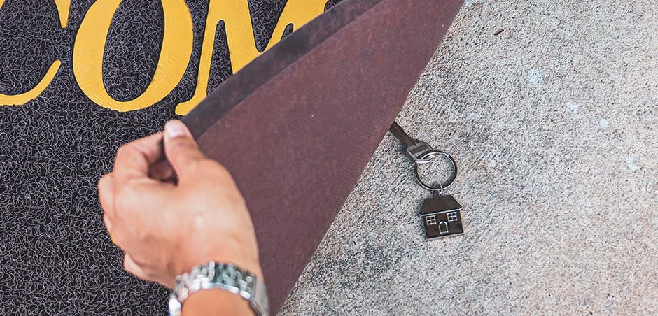 A hand lifts a welcome mat to reveal a house key hidden underneath.