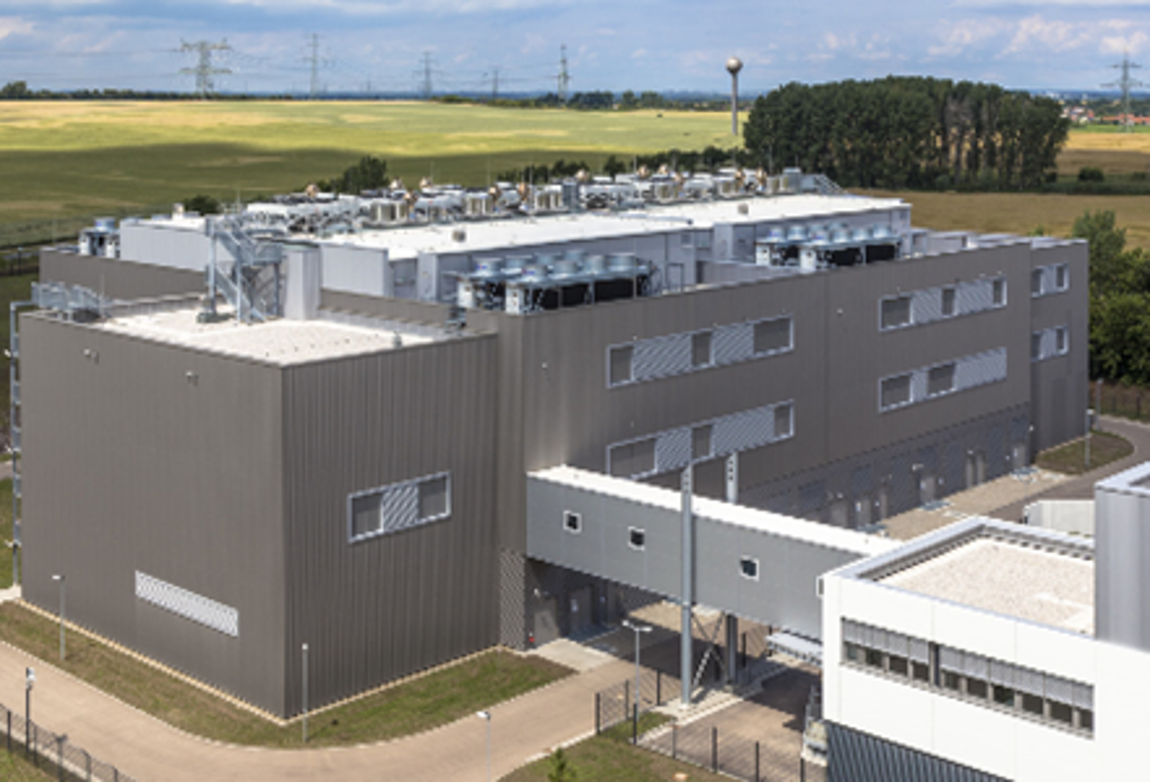 An aerial view of the Twin Core Cloud Data Center in Magdeburg, featuring a large industrial building with rooftop equipment and a connecting skybridge, set in a rural landscape.