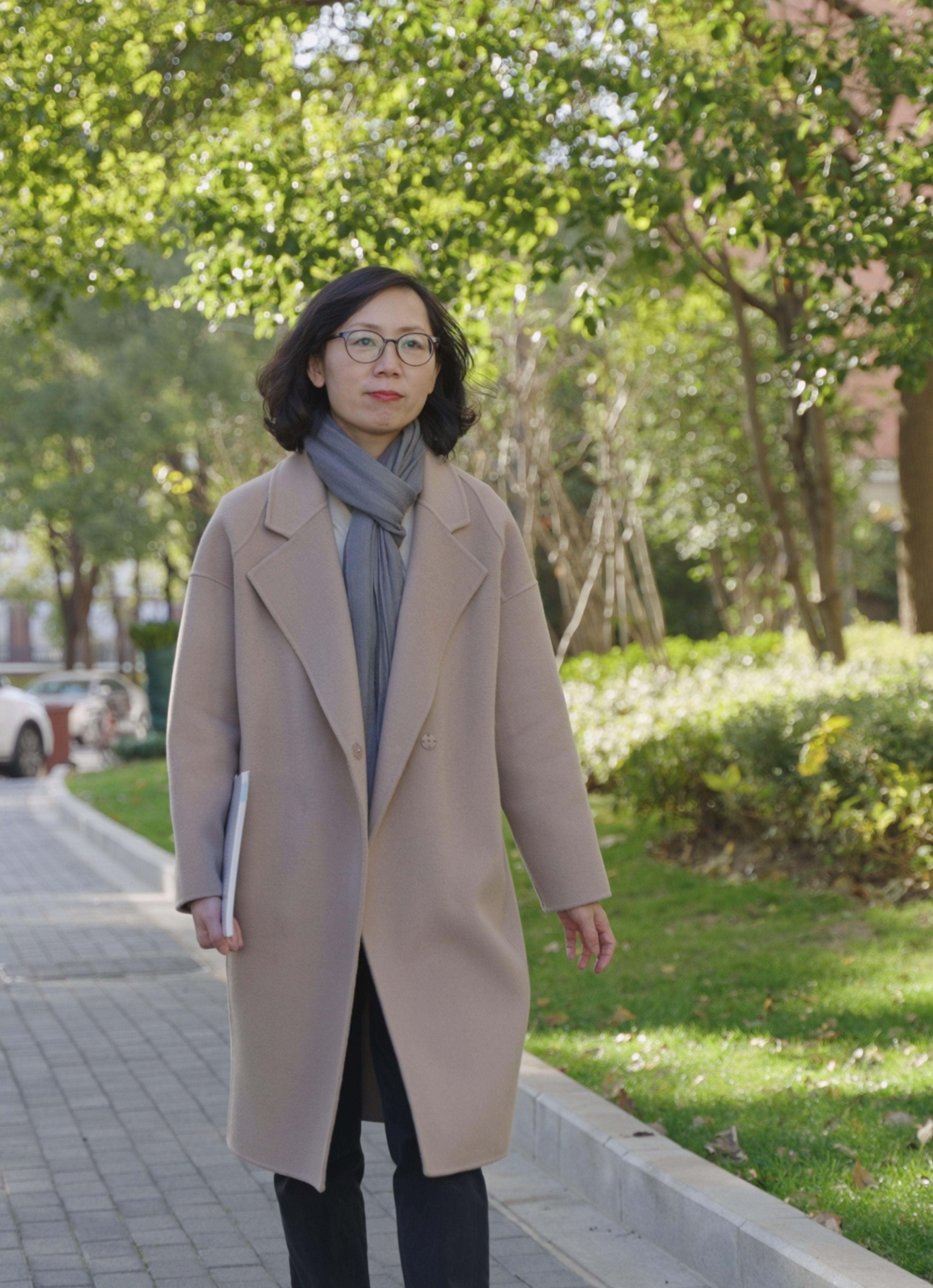 Selina Jiang is walking outdoors in a park, dressed in a beige coat and gray scarf, holding a document.