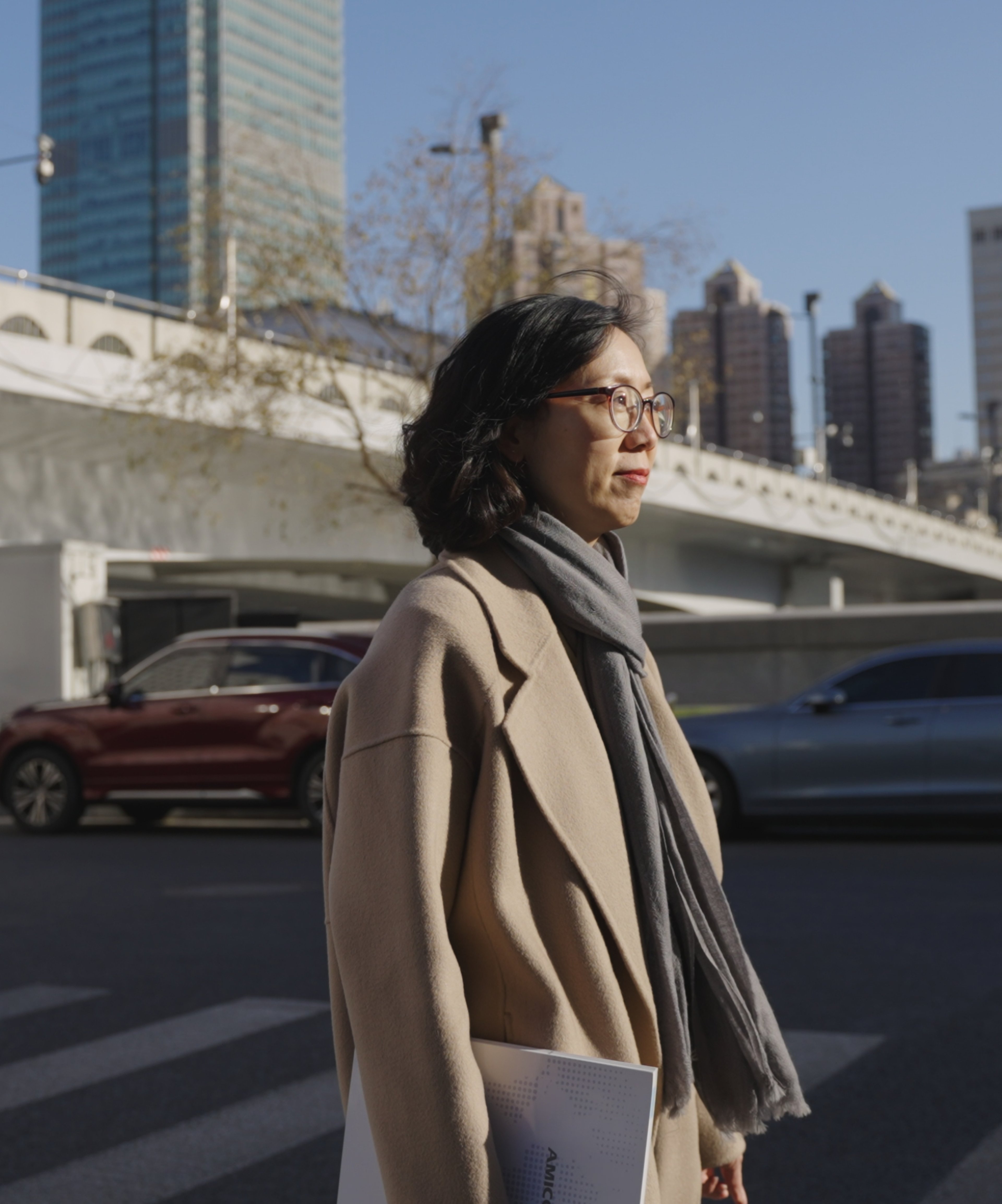 Selina Jiang is standing outdoors in a city, dressed in a beige coat and scarf, holding a document, with modern buildings in the background.