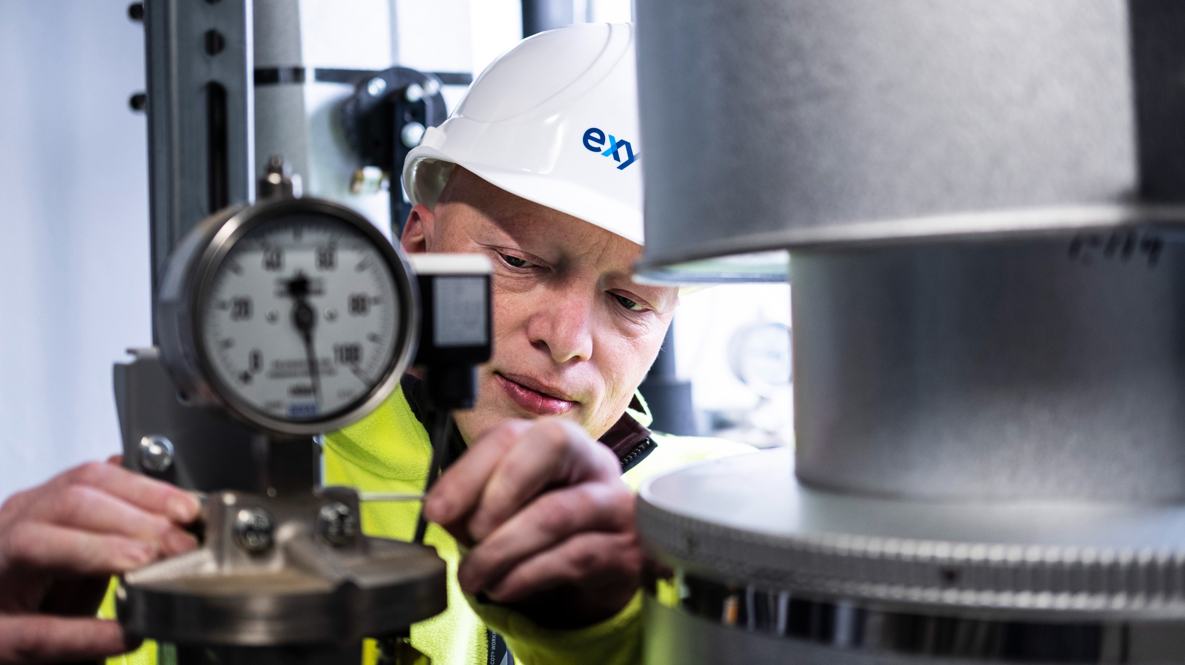 A technician wearing a white Exyte safety helmet and yellow jacket is adjusting a measuring device in an industrial setting.