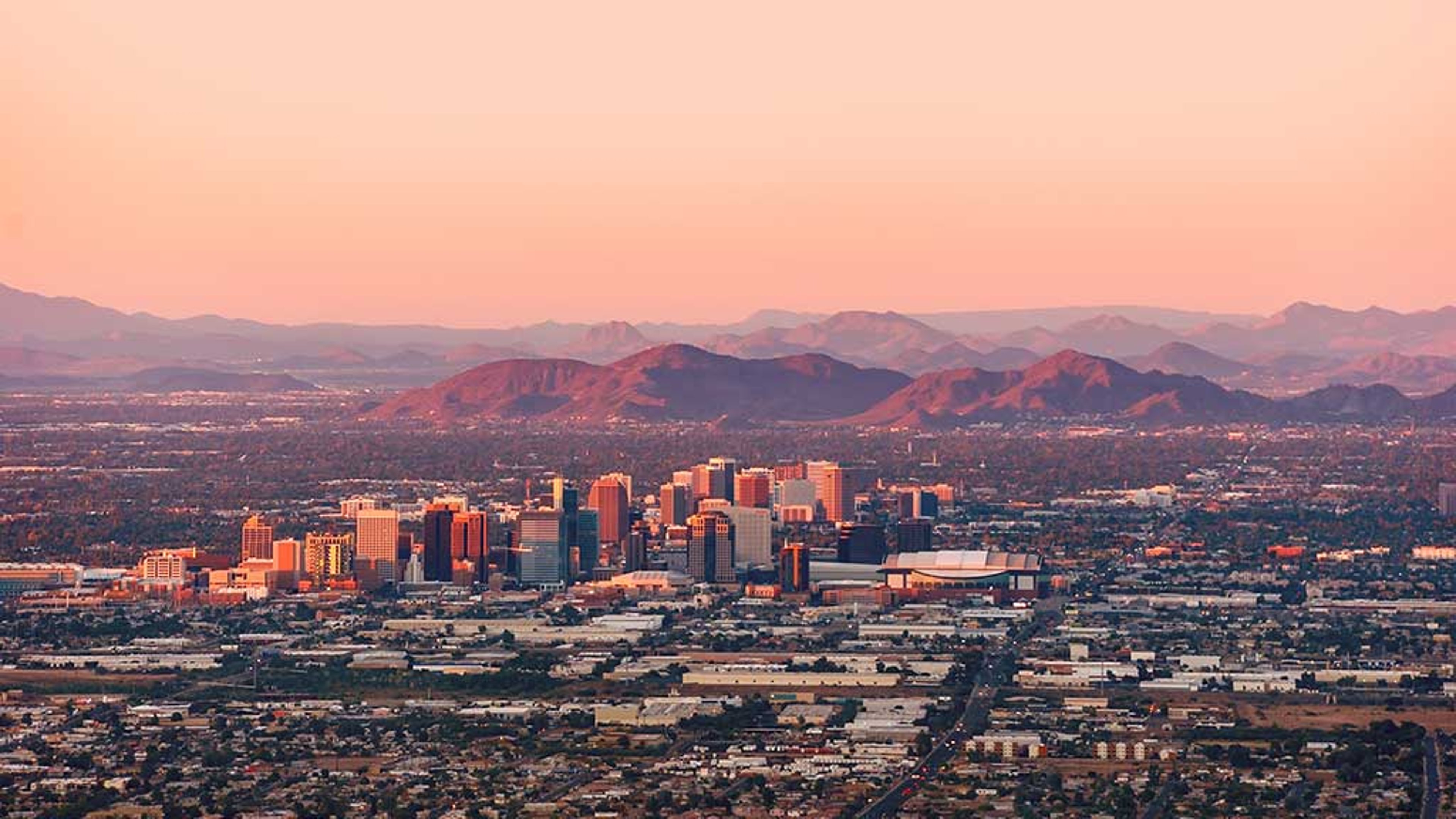 The skyline of Phoenix, Arizona is captured at sunset, with the city buildings bathed in warm light, and the mountains creating a picturesque backdrop under the soft pink sky.