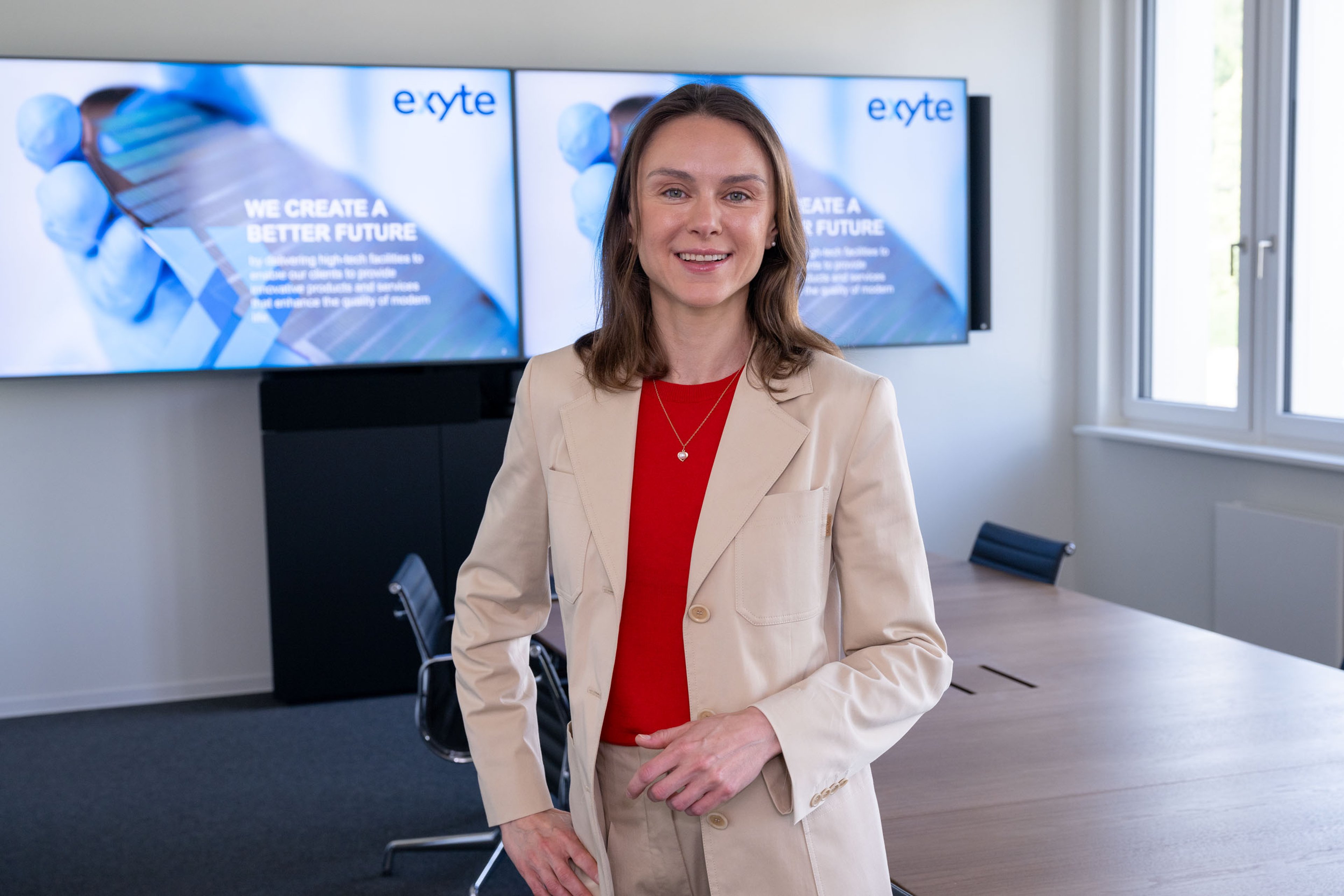 Elena Bashkeeva stands in a modern conference room, dressed in a beige suit with a red top. Behind her, two screens display the Exyte logo and the text “We Create a Better Future,” highlighting the company’s commitment to high-tech solutions.