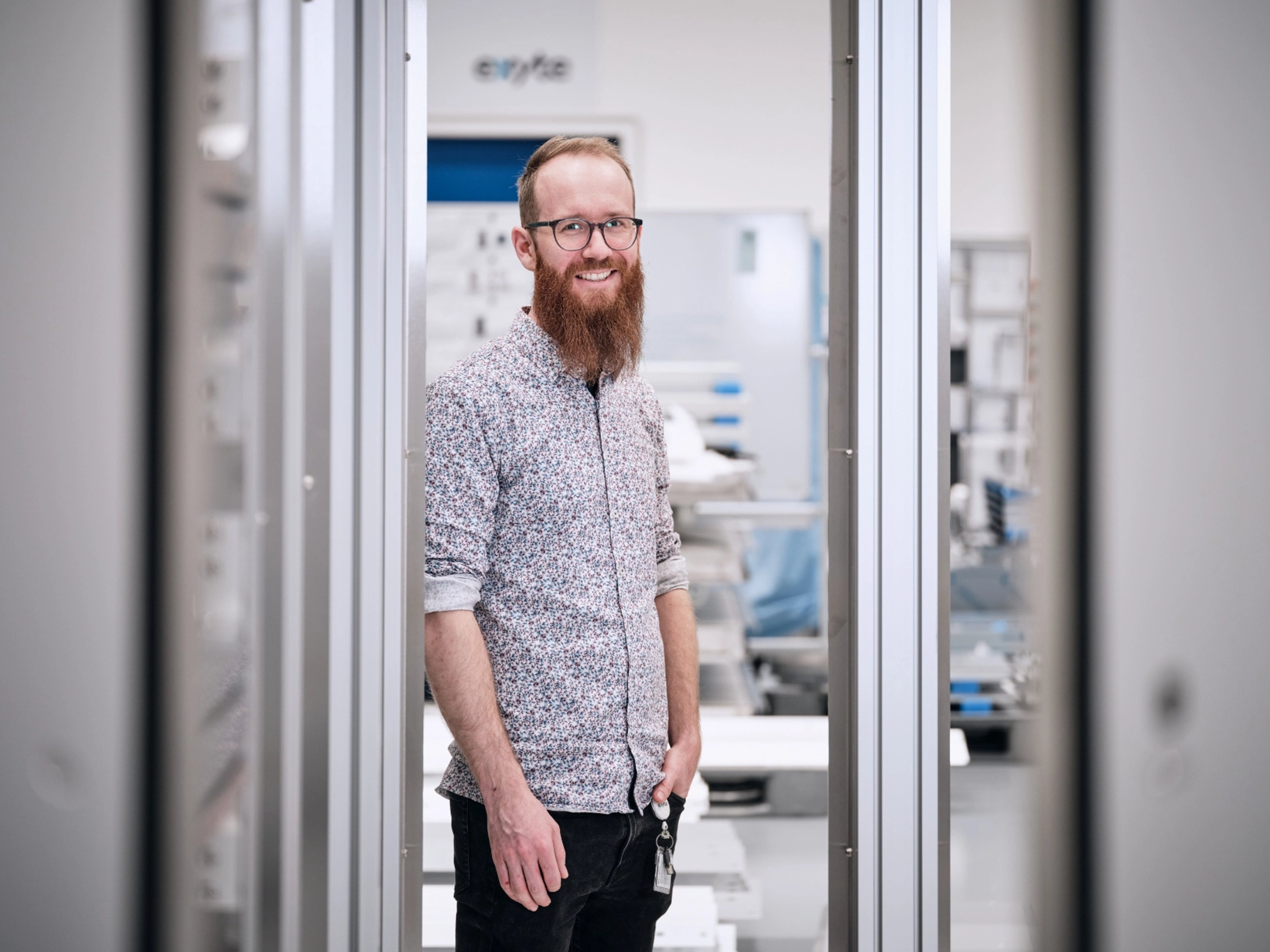Fabian Ankele smiling in a high-tech industrial setting, wearing glasses, a patterned shirt, and sporting a long red beard, standing between two metal frames.