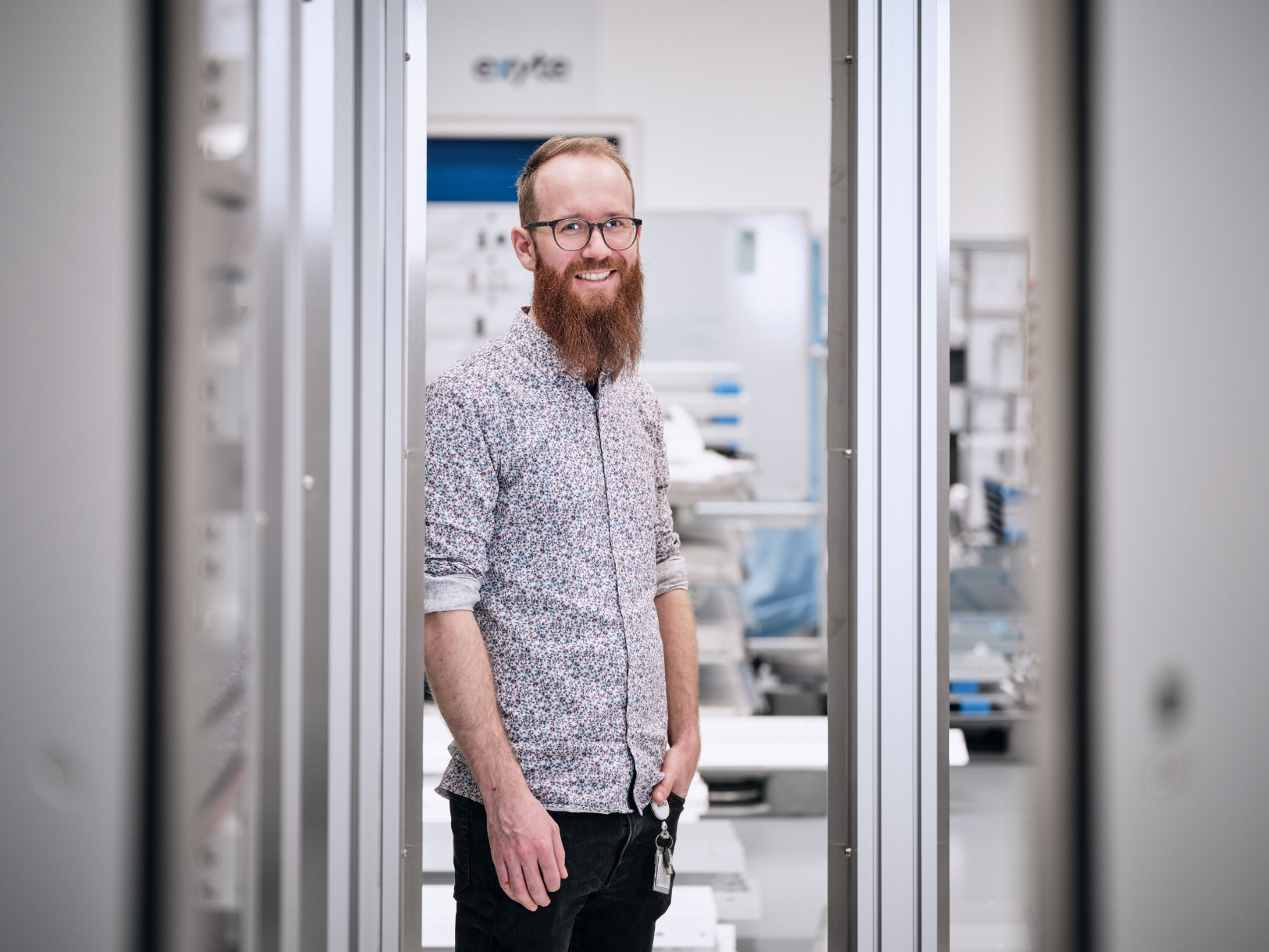 Fabian Ankele smiling in a high-tech industrial setting, wearing glasses, a patterned shirt, and sporting a long red beard, standing between two metal frames.