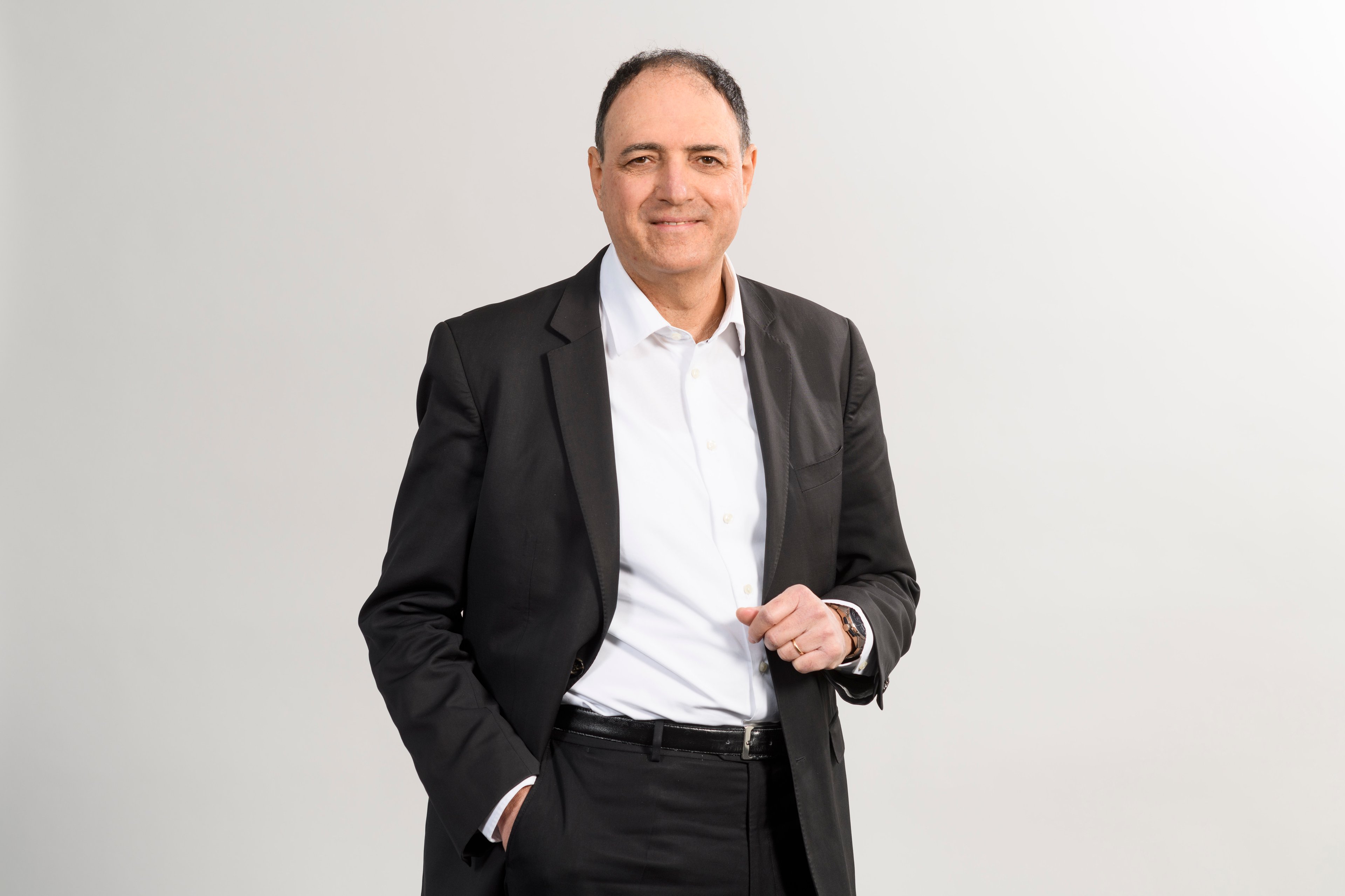A portrait of Luca Mussati, dressed in a black suit and white shirt, standing confidently with one hand in his pocket and the other slightly raised, against a neutral background.
