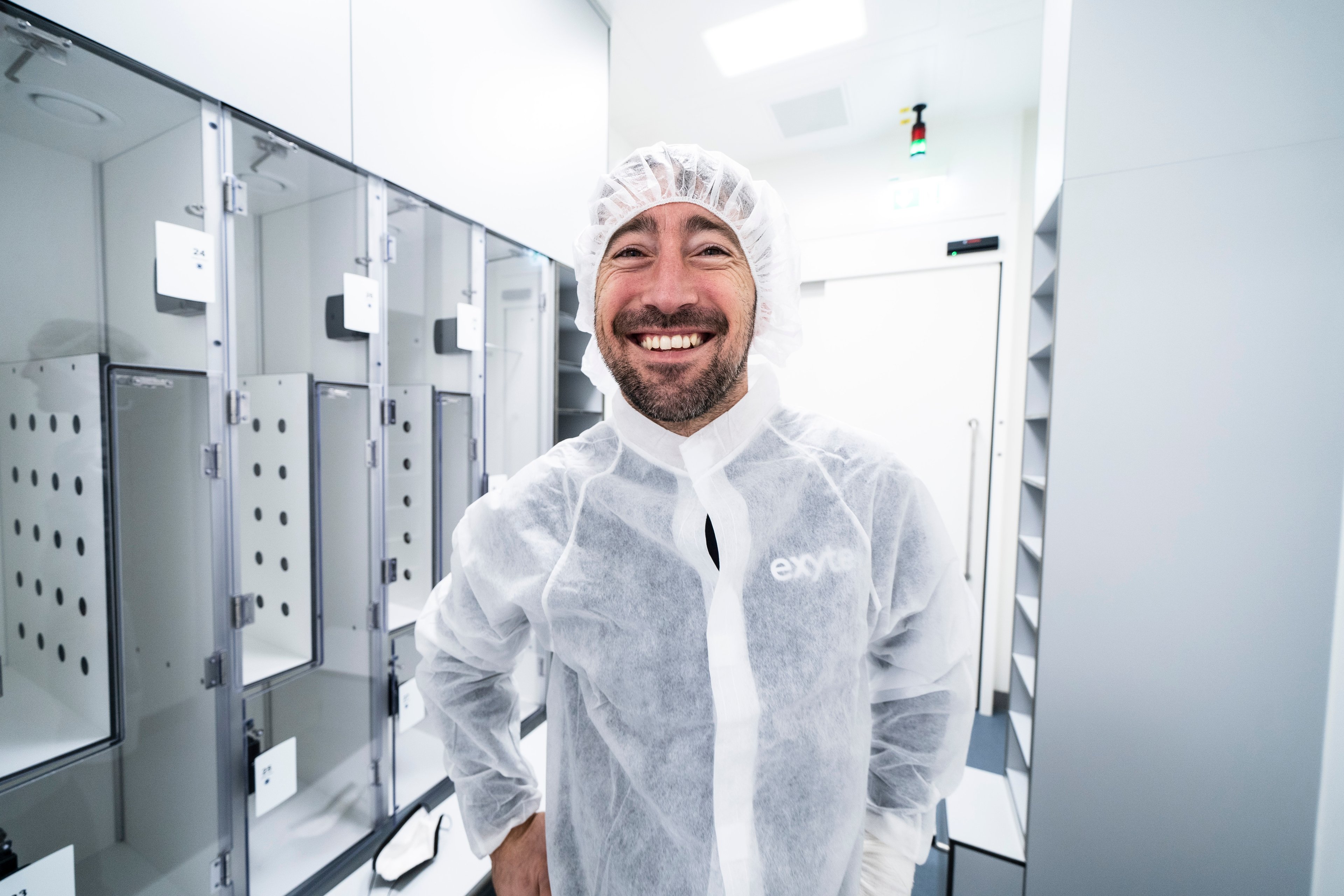 A man in a cleanroom suit smiling in a sterile environment.