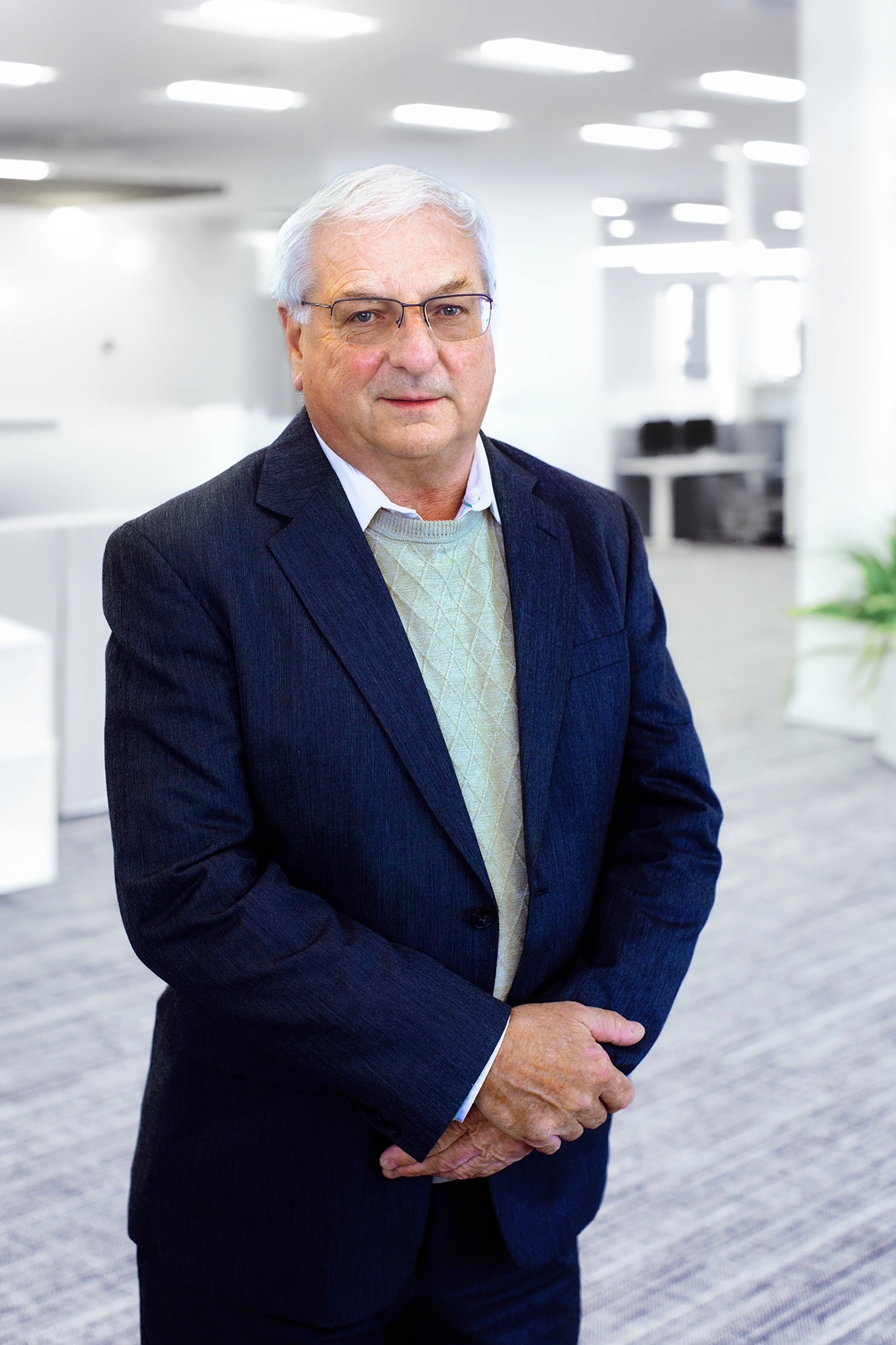 Andrew Whitworth stands in a modern office setting, dressed in a formal suit with a light sweater underneath. His expression is composed, and he is seen with his hands loosely clasped in front.