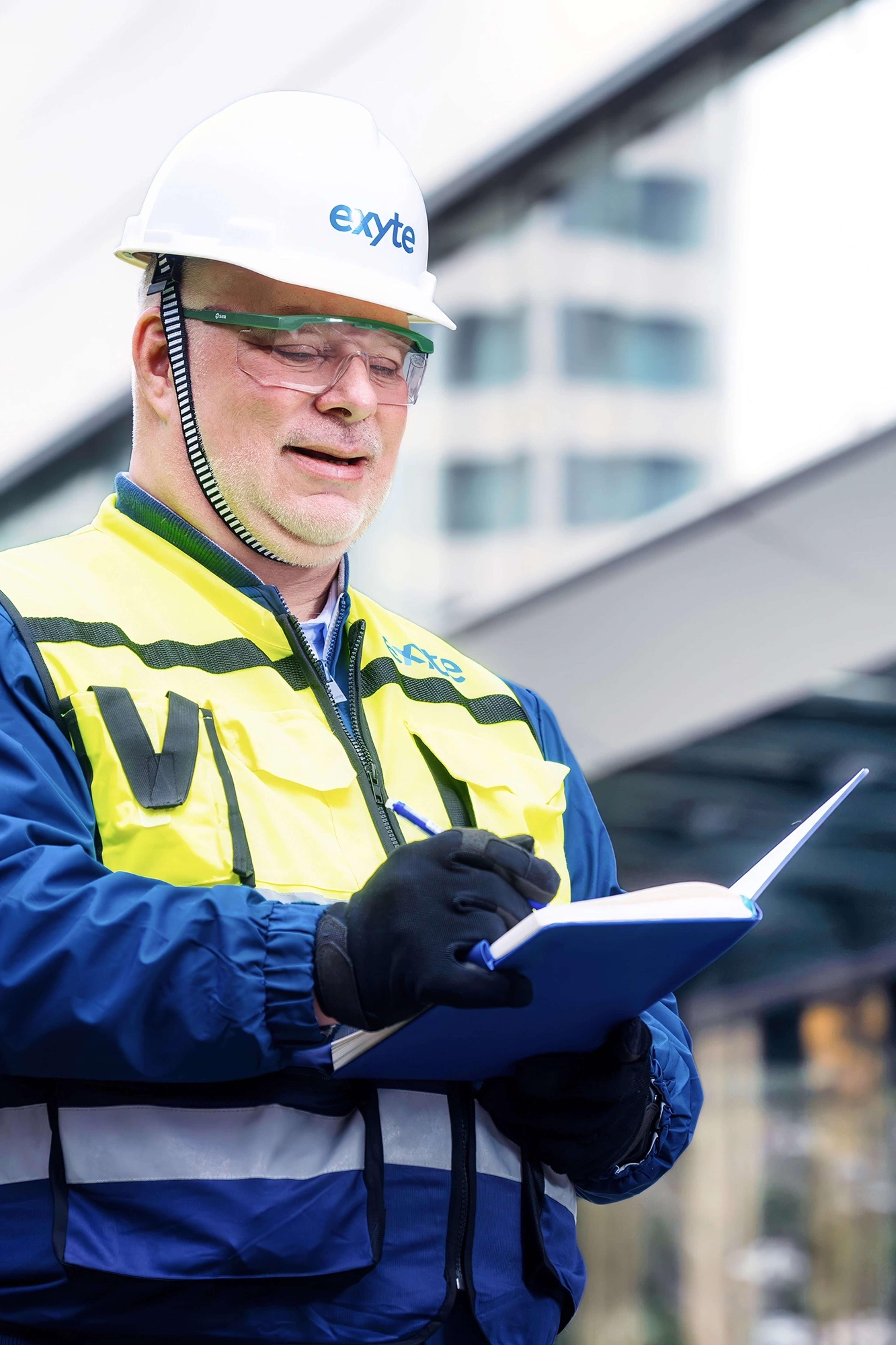 Arend Suhr, wearing safety gear including a hard hat, safety glasses, and gloves, is holding a pen and notebook, appearing to take notes while working outdoors.