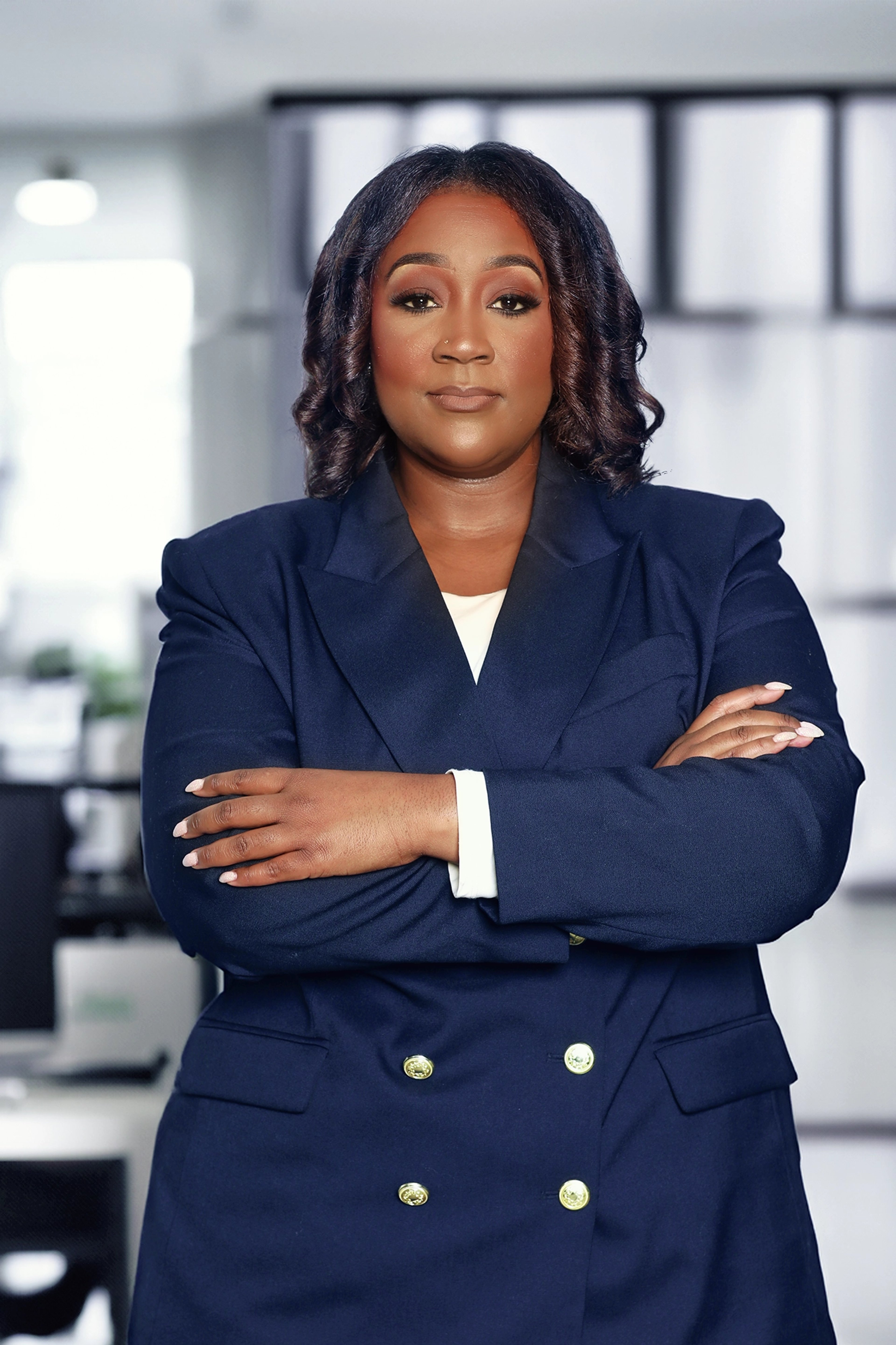 Myesha Daniels stands confidently with arms crossed, dressed in a navy double-breasted blazer. She exudes professionalism in an office setting with her composed expression and poised stance.