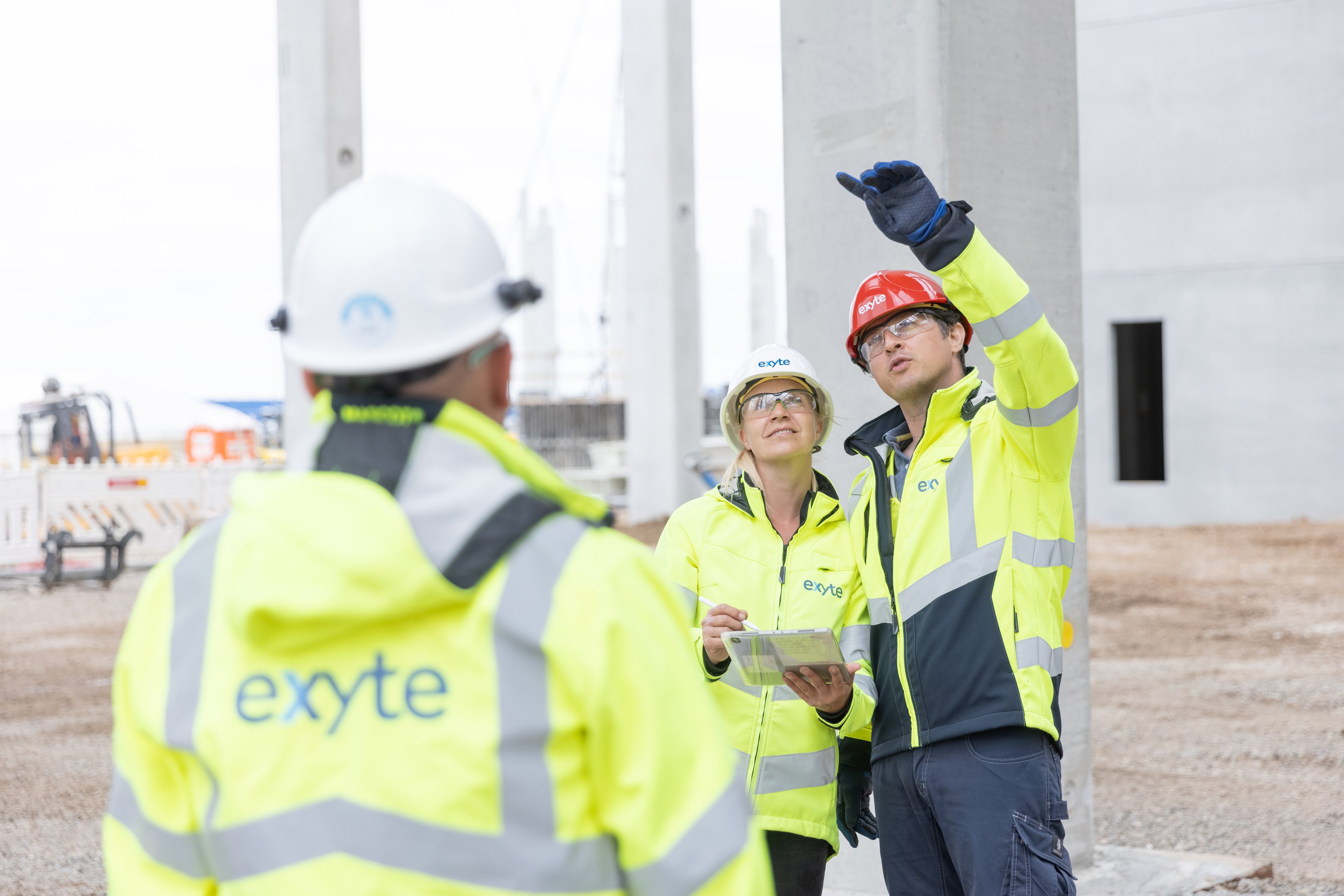 Three construction workers from Exyte, wearing high-visibility jackets and safety helmets, are standing on a construction site. One worker is pointing ahead, while another is holding a clipboard, and the third has their back to the camera.