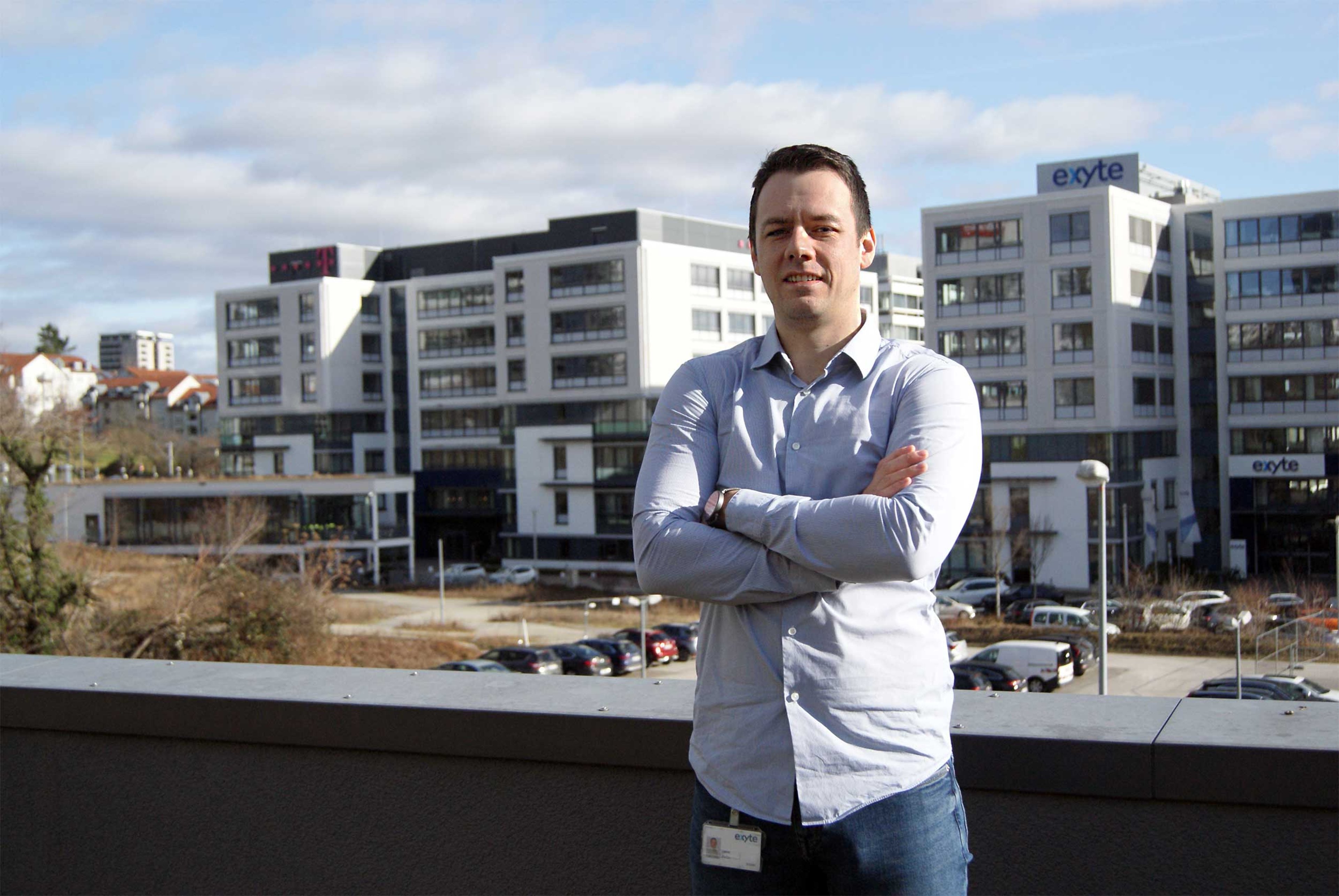 Stefan Ćaćić is standing outside on a balcony with arms crossed, smiling. In the background are modern office buildings with the Exyte logo.