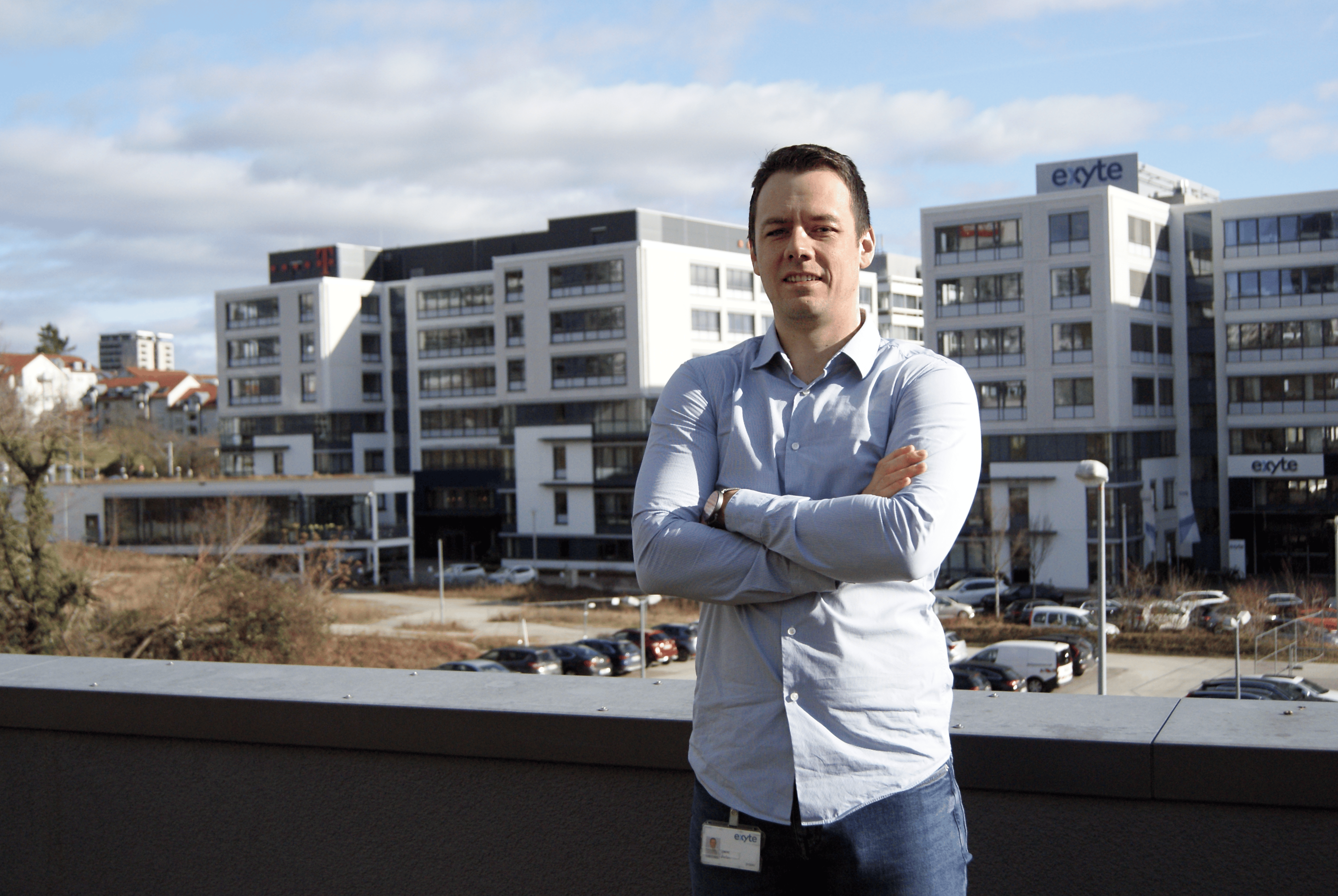 Stefan Ćaćić is standing outside on a balcony with arms crossed, smiling. In the background are modern office buildings with the Exyte logo.