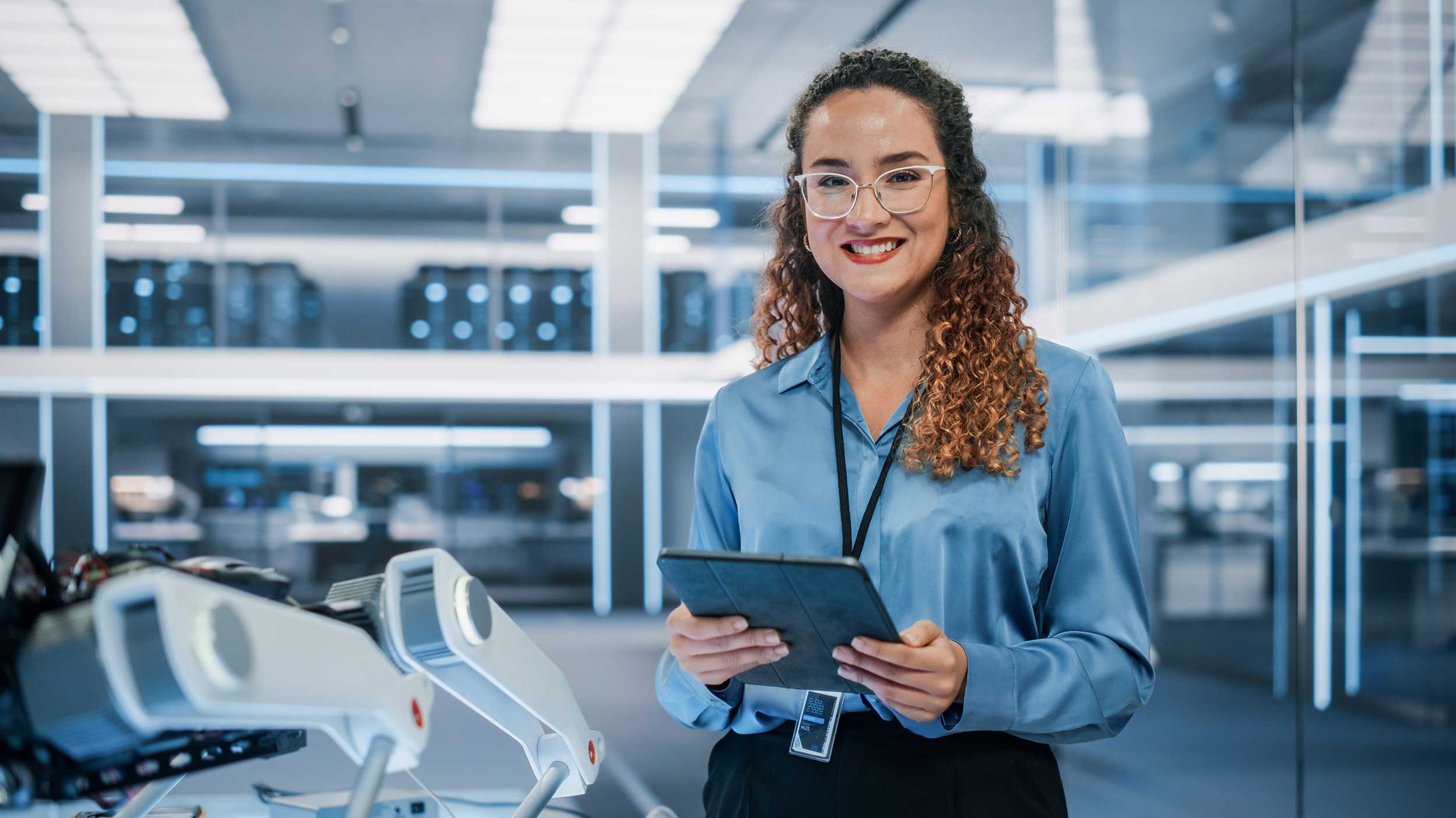 A woman standing in a modern technology lab, smiling while holding a tablet. She is wearing glasses and a blue blouse, with advanced robotic equipment visible in the background.
