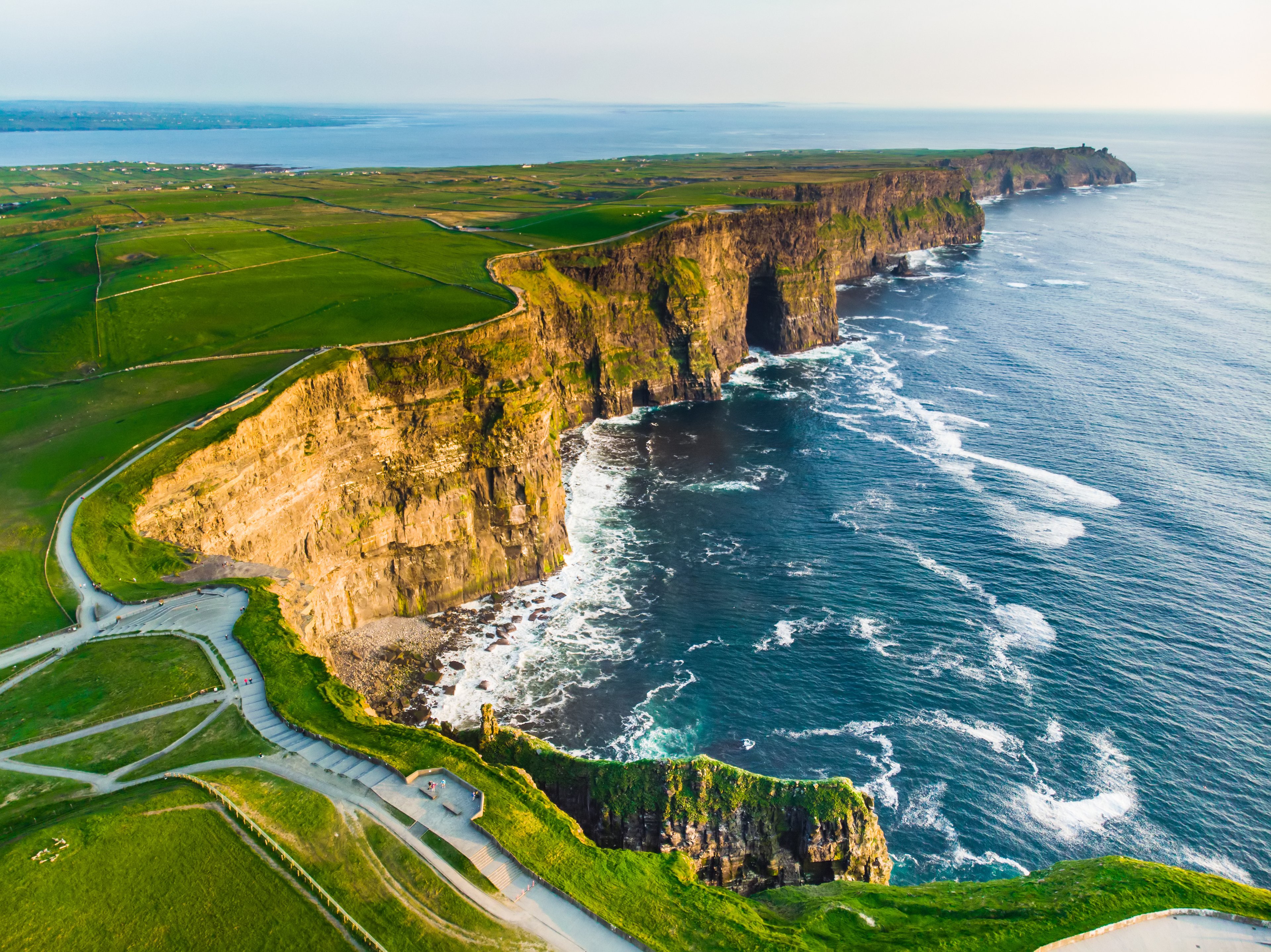 Aerial view of the famous Cliffs of Moher in Ireland. The dramatic cliffs drop steeply into the Atlantic Ocean, with green fields stretching across the top. The sea below is rough, with waves crashing against the cliff face.