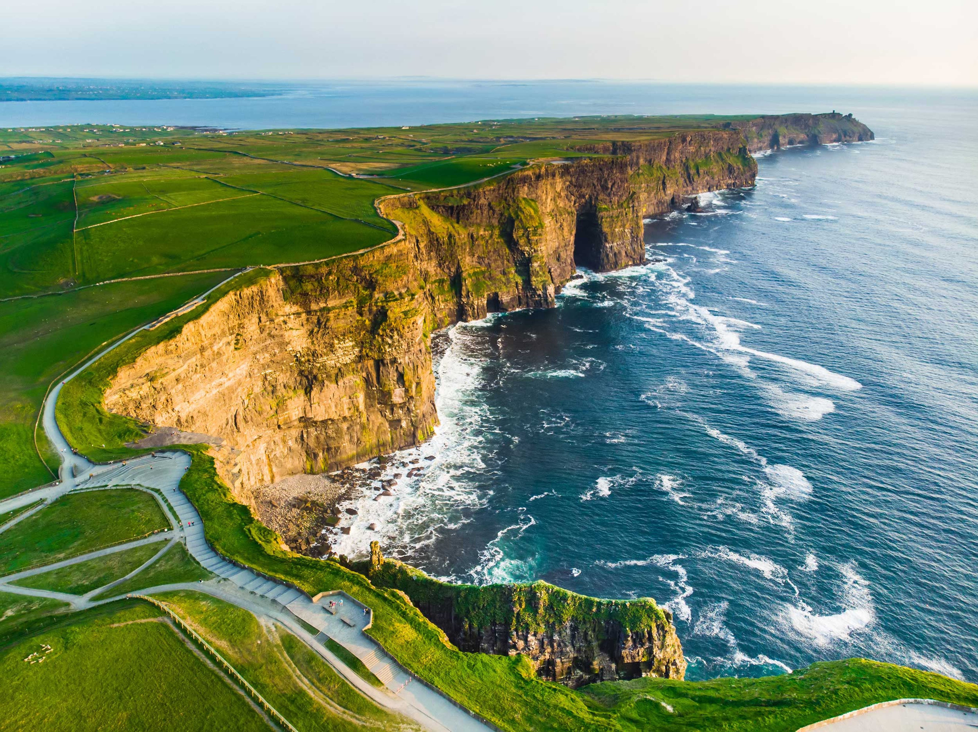 Aerial view of the famous Cliffs of Moher in Ireland. The dramatic cliffs drop steeply into the Atlantic Ocean, with green fields stretching across the top. The sea below is rough, with waves crashing against the cliff face.